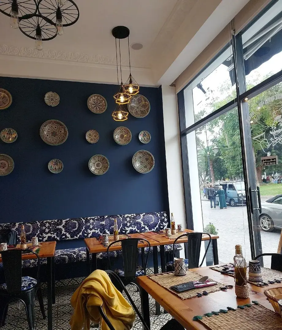 Salle du Restaurant Basidi : mur bleu couvert d'assiettes en faïence, banquette à motifs, baie vitrée donnant sur un jardin.